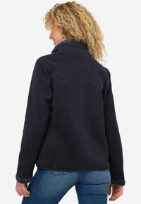 Mörkblå, mjuk tygjacka med hög krage och fluffiga manschetter. Löst skuren, slät textur och enkel design på baksidan. Inga synliga knäppningar eller mönster.