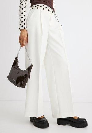 Person iført hvide bukser med vide ben, sorte chunky platformsko, en sort og hvid prikket skjorte og bærende på en lille sort håndtaske med kæderem.