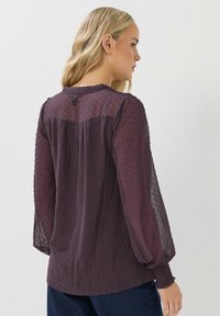 Kobieta z blond włosami nosi przezroczystą, ciemnopurpurową bluzkę z długim rękawem, pokrytą kropeczkami, z marszczonymi mankietami, stoi bokiem na jasnym tle.