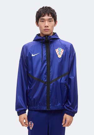 Fiatal férfi kék sportdzsekit és nadrágot visel, melyeken a horvát labdarúgó-válogatott címere és Nike logók láthatók, egy egyszerű háttér előtt áll.