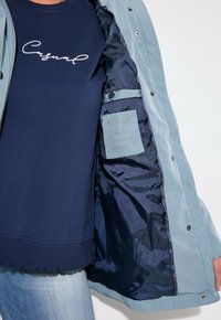 Ljusblå jacka med en marinblå innerfoder, som har slät tyg, tryckknappar och en etikett som indikerar vattentåliga och andningsbara egenskaper.