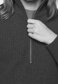 Szary sweter dzianinowy z pół-zamkiem, z teksturą prążkowaną. Ręka chwyta suwak, podkreślając metalowy detal.