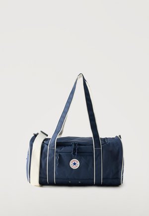 Sac polochon bleu marine avec des bretelles blanches, poche avant zippée et logo circulaire Converse All Star sur fond neutre.