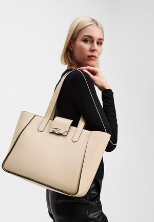 Beige leather tote bag with black trim, featuring two handles and a front closure. Embellished with a silver logo detail. Smooth texture.