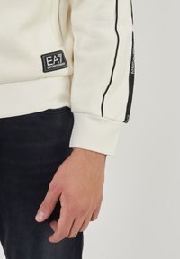 Felpa crema con dettagli neri, caratterizzata da un patch con logo ricamato. Texture liscia con maniche aderenti e cuciture a contrasto.