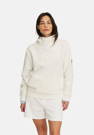 Vrouw met lang donker haar, gekleed in een witte coltrui met logo en witte korte broek, staand tegen een effen achtergrond.