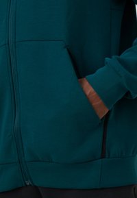 Teal zip-up hoodie featuring a front pocket with a brown accent, ribbed cuffs, and a slim fit. The fabric has a soft, textured finish.