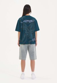Teal oversized t-shirt med ryggrafik, der viser hvid "Ed Hardy" logo og svunget design; parret med lyseblå denimshorts.