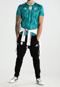 Muž v zeleném dresu německého fotbalu, černých teplácích Adidas, bílých kopačkách a s bílou bundou uvázanou kolem pasu, stojící s rukama v kapsách.