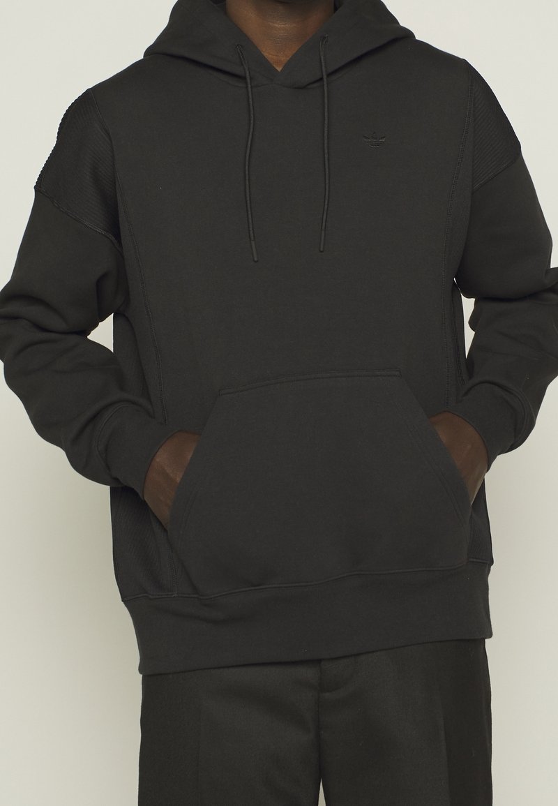 Černá mikina s kapucí vyrobená z měkké tkaniny, s klokaní kapsou, šňůrkami a texturovanými panely na ramenou.