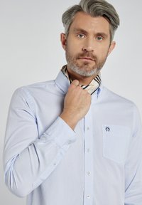 Jasnoniebieska koszula z długim rękawem, z kieszenią po lewej stronie, zdobiona haftowanym emblematem. Mężczyzna zawiązuje wąski szalik w paski wokół szyi.