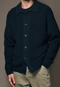 Cardigan en tricot vert foncé avec col et boutons noirs, présentant une texture côtelée et une coupe décontractée. Associé à un pantalon de couleur claire.