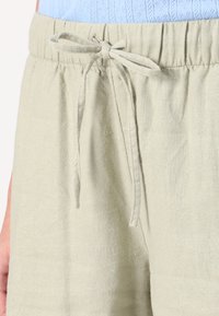 Pantalons en lin vert clair avec taille à cordon, tissu texturé et deux poches latérales. Le design est décontracté et informel.
