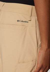 Beige cargo nadrág, amely logóval, külső zsebekkel és a tervezett textúrában látható könnyű anyaggal rendelkezik.