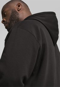 Sudadera con capucha negra de tela texturizada, con un corte relajado y costuras visibles en la capucha y el hombro.