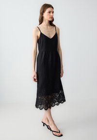 Zwarte mouwloze jurk met een V-hals, een getailleerde bodice en een zoom afgewerkt met kant. De stof heeft een gestructureerde, lichte uitstraling.