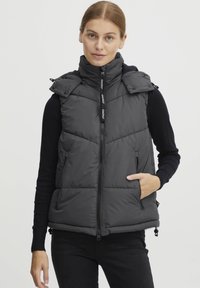 Veste matelassée grise foncée avec un col montant, fermeture éclair devant, deux poches latérales et ourlet ajustable, arborant une texture matelassée et un logo.