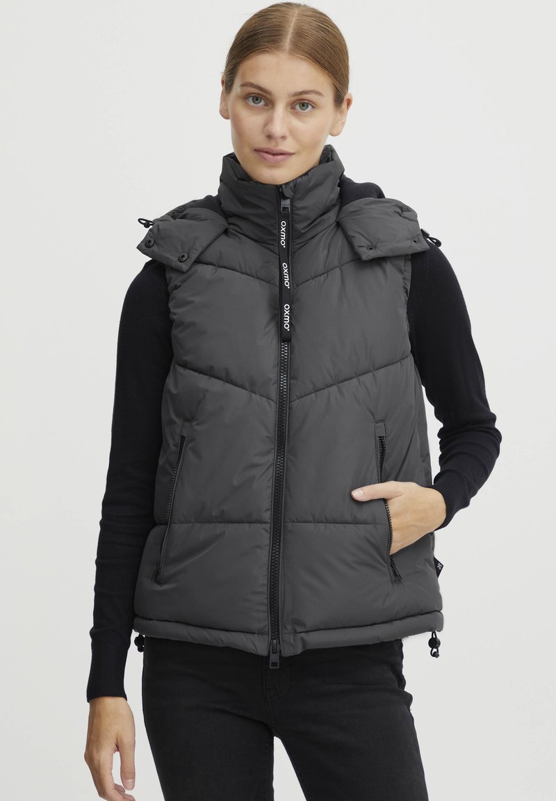 Veste matelassée grise foncée avec un col montant, fermeture éclair devant, deux poches latérales et ourlet ajustable, arborant une texture matelassée et un logo.