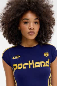 Tânără cu păr creț purtând un tricou bleumarin cu text galben „Portland” și logo-uri ale cluburilor sportive.