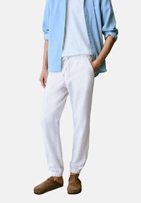 Pantaloni jogger bianchi in lino con polsini elasticizzati, abbinati a una camicia button-up azzurro chiaro e calzature slip-on marroni. Design semplice e dalla vestibilità rilassata.