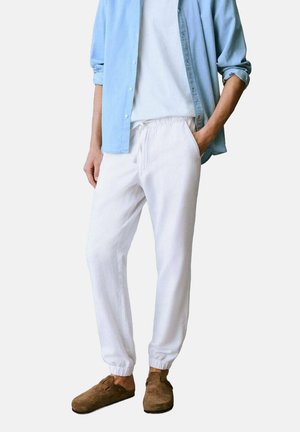 Pantalones jogger de lino blanco con puños elásticos, combinados con una camisa de botón de color azul claro y calzado marrón sin cordones. Diseño simple y de corte relajado.