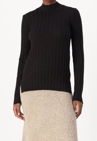 Maglione lungo a maniche lunghe nero a coste con collo alto, silhouette attillata e tessuto testurizzato; indossato sopra una gonna beige in peluche.