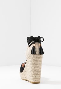 Espadrille noire à talon en forme de coin avec un tissu beige tissé, présentant un talon en jute tressé, des lacets noirs et des accents de gland au niveau de la cheville.