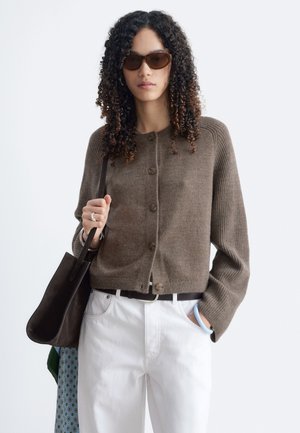 Femme aux cheveux bouclés portant un cardigan marron, un pantalon blanc, des lunettes de soleil, portant un sac à bandoulière sombre et une écharpe à motifs.
