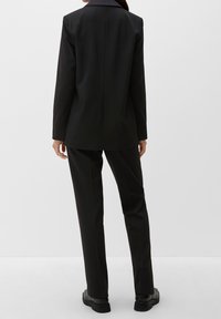 Blazer nero su misura con revers, abbinato a pantaloni coordinati. Il tessuto è liscio con dettagli minimi e una vestibilità dritta.