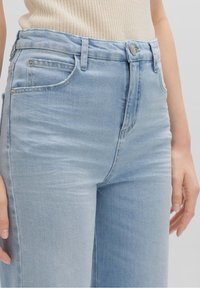 Jeans de tiro alto de color azul claro combinados con una blusa sin mangas de punto beige, que muestra la cintura, las caderas y las manos.