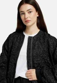 Svart bomberjacka med texturerad tyg, ribbade ärmslut och en silverfärgad dragkedja. Bärs över en vit t-shirt, som har en avslappnad passform och voluminösa ärmar.