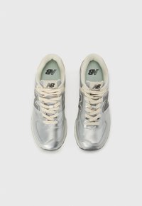 Zapatillas deportivas plateadas con una parte superior sintética suave, cordones beige y un cuello ligeramente acolchado. Presentan un destacado logo "N" en gris oscuro.