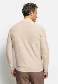 Beige gestreifter Pullover mit langen Ärmeln und gerippten Bündchen; besitzt einen runden Ausschnitt. Stricktextur, figurbetonte Silhouette, getragen mit braunen Hosen.