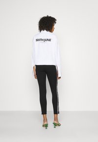 Camicia oversize bianca a righe con logo "SIXTH JUNE", abbinata a leggings neri attillati con righe bianche laterali e sandali con tacco verdi.