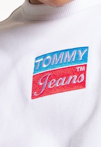Bílá mikina s výšivkou loga "TOMMY" v modré barvě a "Jeans" v červené, s hladkým texturem a žebrovaným výstřihem.