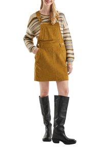 Geel corduroy tuinbroek jurk met een voorzak, gecombineerd met een gebreide trui met patroon. Zwarte kniehoge laarzen met een chunky hak.