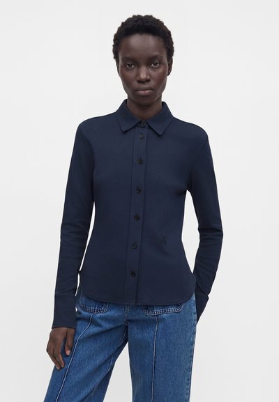 Femme portant une chemise bleu marine à manches longues avec un col et un jean bleu, debout devant un fond clair uni.