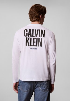 Uomo che indossa una camicia bianca a maniche lunghe con grande testo nero "CALVIN KLEIN" e "ESTABLISHED 1968" sulla schiena, abbinata a jeans blu.
