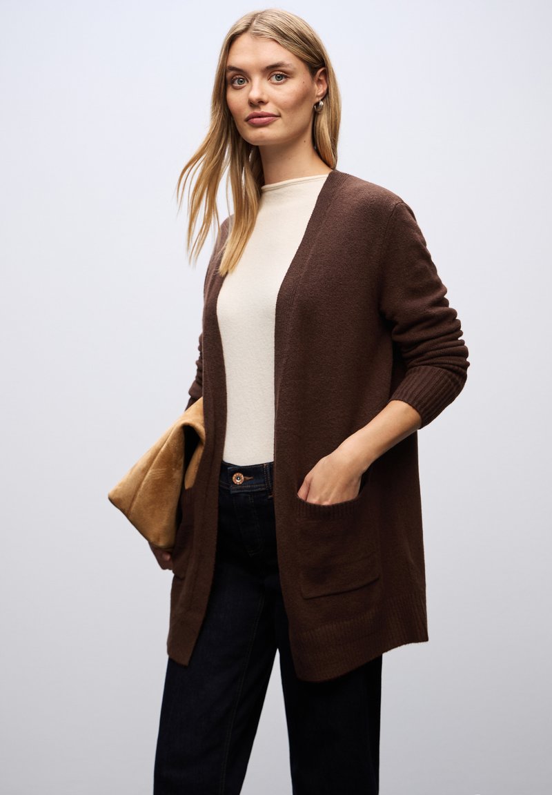 Brown cardigan with open front, ribbed cuffs, and pockets, worn over a light-colored top. Accessory in hand is a tan pouch.