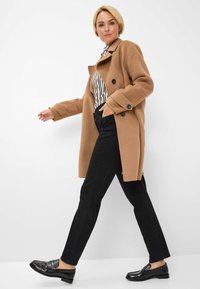 Cappotto in misto cammello con design doppiopetto, bottoni neri, abbinato a jeans neri slim e mocassini neri, con una maglietta a righe.