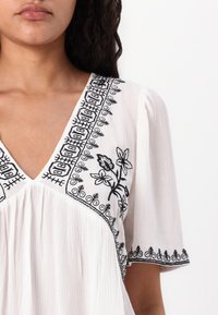 Hvid bluse med V-udskæring og korte ærmer, der har sort blomsterbroderi og intrikate kantmønstre langs udskæringen og ærmerne.