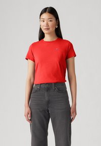 Rotes Baumwoll-T-Shirt mit kurzen Ärmeln, Rundhalsausschnitt, kleinem Logo auf der Brust, kombiniert mit hochgeschnittenen grauen Jeans.