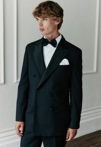 Schwarzer, doppelreihiger Smoking-Jacket mit satinierten Revers, weißem Hemd und schwarzer Fliege. Verfügt über ein weißes Einstecktuch und eine taillierte Passform.