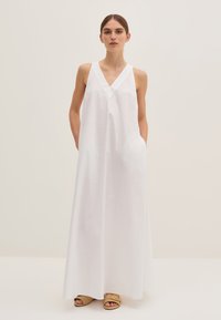 Abito maxi bianco senza maniche con scollatura a V, realizzato in tessuto liscio. Presenta tasche laterali e una vestibilità comoda. Indossato con sandali beige.