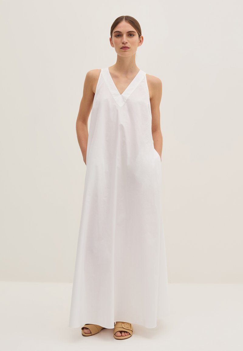 Abito maxi bianco senza maniche con scollatura a V, realizzato in tessuto liscio. Presenta tasche laterali e una vestibilità comoda. Indossato con sandali beige.