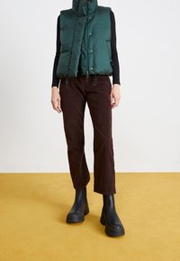 Chaleco verde acolchado con cuello alto, camiseta de manga larga negra, pantalones de pana marrones y botas negras de suela gruesa contra un fondo amarillo.