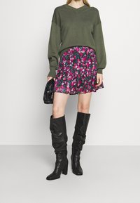 Pull oversize vert olive, jupe courte fleurie rose et noire, et bottes à talons en cuir noir montantes jusqu'aux genoux. Sac à main noir en texture.