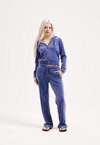 Világoskék velúr melegítő, cipzáros, rövidített kapucnis pulóverrel, húzózsinórokkal és hozzáillő melegítőnadrággal, oldalzsebekkel és rugalmas derékrésszel.