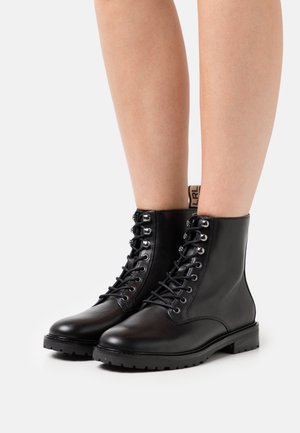 Nilkat ja alaraajat, joissa mustat nahkaiset nyörilliset taistelukengät paksuilla kumipohjilla valkoisella taustalla.