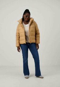 Γυναίκα φόρεσε μπεζ μπουφάν, λευκό πουλόβερ, μπλε τζιν, γκρι αθλητικά παπούτσια και μπεζ γυαλιά ηλίου, στέκεται σε απλή ανοιχτόχρωμη φόντο.